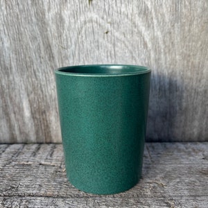 May include: A dark green ceramic planter with a speckled finish. The planter is cylindrical in shape and has a smooth, rounded rim.