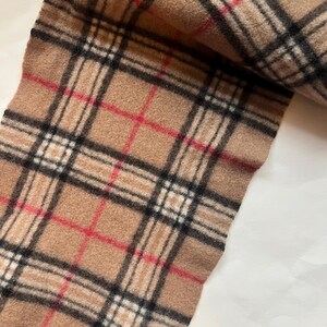 May include: A tan scarf with a classic plaid pattern in black, red, and white. The scarf has a fringed edge and is made of a soft, woven material. This accessory is suitable for cold weather.