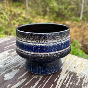 Puede incluir: Una maceta de cerámica con exterior azul oscuro y negro, con dos rayas blancas horizontales. La maceta tiene una base de pedestal y una abertura ancha. La maceta está sobre una superficie de madera desgastada.