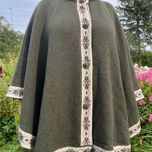 Könnte beinhalten: Ein grüner, gestrickter Kapuzenumhang mit einem weißen und braunen Muster am Saum. Der Umhang hat einen Knopfverschluss an der Vorderseite.