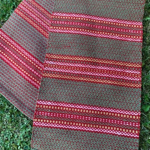 May include: A woven table runner with a green, red, and yellow striped pattern. The runner is made of a textured fabric and has a fringed edge.