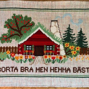 Może przedstawiać: Hafciarski obraz przedstawiający czerwony domek z białymi ozdobami i zielonymi drzewami. Domek ma dwa okna i drzwi. Pod domkiem wyszyty jest napis "BORTA BRA MEN HEMMA BAST".