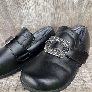 Könnte beinhalten: Schwarze Lederschuhe mit dekorativen silbernen Schnallen. Die Schuhe haben ein klassisches Design mit runder Spitze und einem Riemen über dem Oberteil. Die Schnallen sind verziert und verleihen dem Schuhwerk einen Hauch von Eleganz.