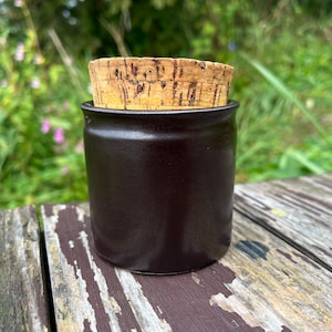 Op de afbeelding: Een donkerbruine keramische pot met een kurken deksel. De pot staat op een houten oppervlak.
