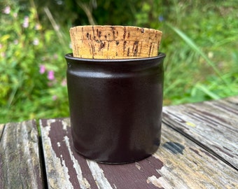 May include: A dark brown ceramic jar with a cork lid. The jar is sitting on a wooden surface.