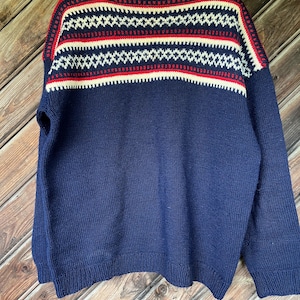 May include: A navy blue sweater with a patterned yoke. The yoke features a repeating geometric design in white and red. The sweater is made of a thick knit material and has long sleeves. The sweater is hanging on a wooden hanger.