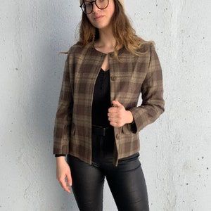 Può includere: Un blazer a quadri marroni e beige con una chiusura a un solo bottone. Il blazer ha una scollatura rotonda e due tasche. La modella indossa pantaloni neri e un berretto beige.