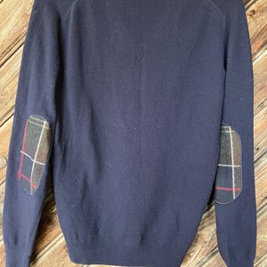 May include: A navy blue sweater with plaid elbow patches. The sweater has a crew neck and long sleeves.