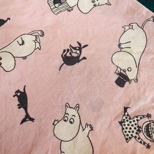 May include: Pink fabric with a repeating pattern of Moomin characters in black and white. The characters are in various poses, including standing, lying down, and jumping. The fabric has a soft, slightly textured appearance.