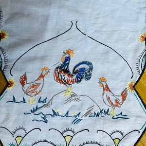 Op de afbeelding: Geborduurde witte tafelloper met drie hanen in oranje, blauw en bruin. De loper heeft een geschulpte rand met bloemen en geometrische ontwerpen in geel, groen en zwart. De hanen zijn geborduurd op een lichtgrijze achtergrond.