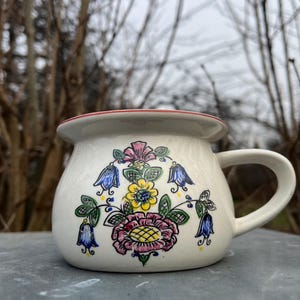 Puede incluir: Taza de cerámica blanca con un borde rojo y un diseño floral en azul, amarillo, verde y rosa. Las flores son estilizadas y se asemejan a campanas.