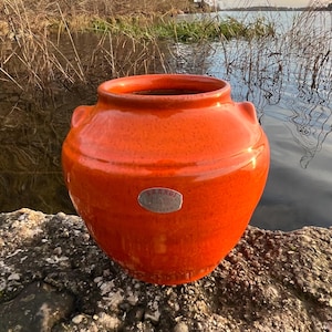 Könnte beinhalten: Ein leuchtend orangefarbener Keramiktopf mit glänzendem Finish. Der Topf hat eine runde Form mit zwei kleinen Griffen und einem kleinen Metall-Emblem. Der Topf steht auf einem Felsen in der Nähe eines Gewässers.
