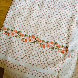 以下が含まれることがあります： 小さなピンクの花が繰り返し描かれた白い布地。大きなピンクの花と緑の葉が縁取りになっている。