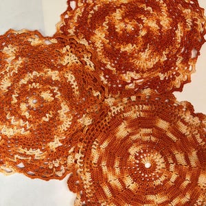 May include: Three crocheted doilies in shades of orange and cream. The doilies are circular and have a delicate, lacy design.
