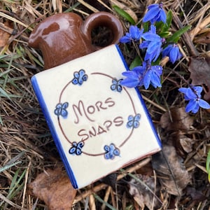 Op de afbeelding: Een kleine bruine keramische kan met een blauw-wit ontwerp. De kan heeft een vierkante basis en een ronde handgreep. De kan is versierd met blauwe bloemen en de woorden "Mors Snaps".