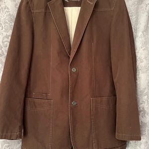 May include: A brown blazer with two front pockets and a two-button closure. The blazer has a notched lapel and a light-colored lining. The word "TRAVEL" is visible on the label.