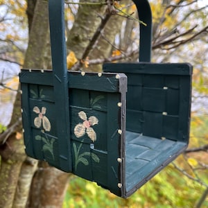 Puede incluir: Una cesta de madera verde oscuro con asa, pintada con puntos blancos y flores rosas. La cesta está colgada de una rama de árbol.