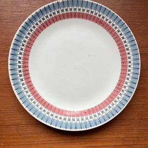 May include: A white ceramic plate with a decorative border. The border features a pattern of blue and red lines with black accents. The plate is round and sits on a wooden surface. This plate is suitable for serving food.