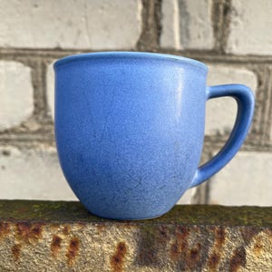 Op de afbeelding: Een blauwe keramische mok met een gebogen oor. De mok heeft een getextureerd oppervlak en een licht uitlopende rand. De mok is effen blauw van kleur en lijkt handgemaakt. De achtergrond is een verweerde bakstenen muur.