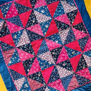 Op de afbeelding: Een handgemaakte quilt met een geometrisch patroon van driehoeken in roze, marineblauw en bloemenprints. De quilt is omrand met een effen marineblauwe stof, wat een opvallend contrast creëert. De quilt heeft een vierkante vorm.