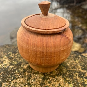 Könnte beinhalten: Eine kleine, hölzerne Dose mit Deckel und einem Knopf oben. Die Dose ist aus hellbraunem Holz und hat eine glatte, polierte Oberfläche.