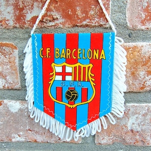 Puede incluir: Un banderín con el logotipo del FC Barcelona, con fondo azul y rayas verticales rojas. El banderín tiene una franja blanca y un cordón blanco para colgarlo. El escudo del equipo está en el centro, con el texto "C.F. Barcelona".
