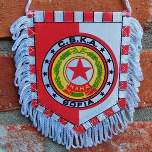 Puede incluir: Un banderín con diseño rojo, blanco y dorado. Presenta un emblema central con una estrella roja y el texto "C.S.K.A. SOFIA". Flecos blancos adornan los bordes y una cuerda blanca está adjunta para colgar.