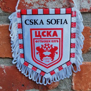 Puede incluir: Pequeño banderín con el texto "CSKA SOFIA" y un escudo del equipo. El banderín es blanco con detalles rojos y negros, y flecos blancos. El escudo presenta un escudo con un balón de fútbol y dos osos rojos.