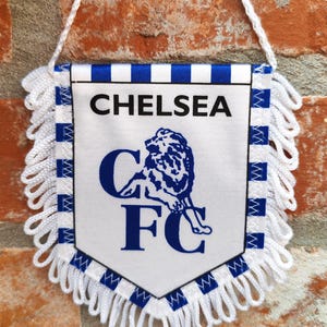 Puede incluir: Bandera blanca con ribete azul y blanco, con el texto "CHELSEA" y un emblema de león azul. La bandera está colgada por un cordón blanco contra una pared de ladrillo.