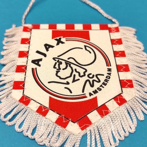 Könnte beinhalten: Ein weiß-rotes Wimpel mit weißem Fransenbesatz. Der Wimpel zeigt das Logo von Ajax Amsterdam, das ein Schwarzweißbild eines Mannes mit Hut enthält. Der Text "AJAX" und "AMSTERDAM" ist in Schwarz gedruckt.