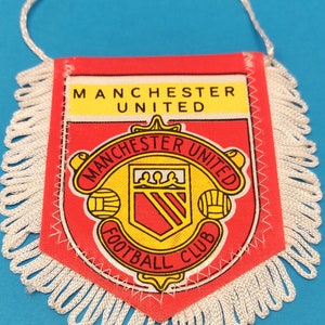 Op de afbeelding: Een rode vlaggetje met de tekst "MANCHESTER UNITED" in het geel. In het midden staat het clublogo, een schild met een geel en rood ontwerp en de tekst "FOOTBALL CLUB". Het vlaggetje heeft witte franjes en een wit touwtje om op te hangen.