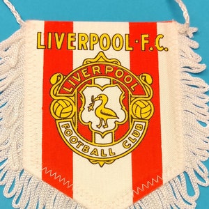 Könnte beinhalten: Wimpel mit dem Logo des Liverpool F.C., mit roten und weißen Streifen und einem weißen Fransenbesatz. Das Logo zeigt einen gelben Vogel, die Worte "Liverpool Football Club" und zwei gelbe Fußbälle.
