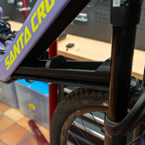 Könnte beinhalten: Ein schwarzer Fahrradrahmen mit dem gelben Text "SANTA CRUZ" auf einem lila Rahmen. Der Vorderreifen ist sichtbar, zusammen mit der Vorderradgabel. Das Fahrrad ist auf einem schwarzen Ständer montiert.