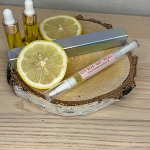Può includere: Un flacone di vetro trasparente con un tappo bianco e un'etichetta rosa su cui è scritto "CIT-RUS GOT REAL! Lemon Sourced Nourishing Oil". Il flacone è appoggiato su una superficie di legno con due limoni e una scatola argentata.