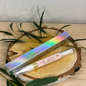 Puede incluir: Un tubo transparente con una tapa blanca que contiene una etiqueta rosa y blanca que dice "BARE WITH ME Aceite nutritivo sin perfume". El tubo está sobre una superficie de madera con hojas verdes.