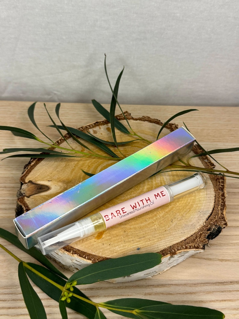 Puede incluir: Un tubo transparente con una tapa blanca que contiene una etiqueta rosa y blanca que dice "BARE WITH ME Aceite nutritivo sin perfume". El tubo est&aacute; sobre una superficie de madera con hojas verdes.