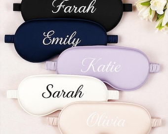 May include: Five personalized sleep masks in black, navy, lavender, white, and blush pink. Each mask has a different name in elegant white script: Farah, Emily, Katie, Sarah, and Olivia. The masks are arranged on a light pink surface.