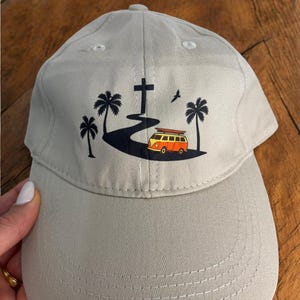 May include: A light grey baseball cap with a graphic of a cross, a road, palm trees, and a vintage orange and yellow van. The cap has a curved peak and a button on top. The design is printed on the front of the cap.
