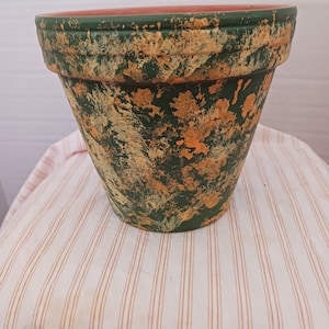 May include: A terracotta flower pot with a green and orange speckled paint finish. The pot is sitting on a white and brown striped surface.