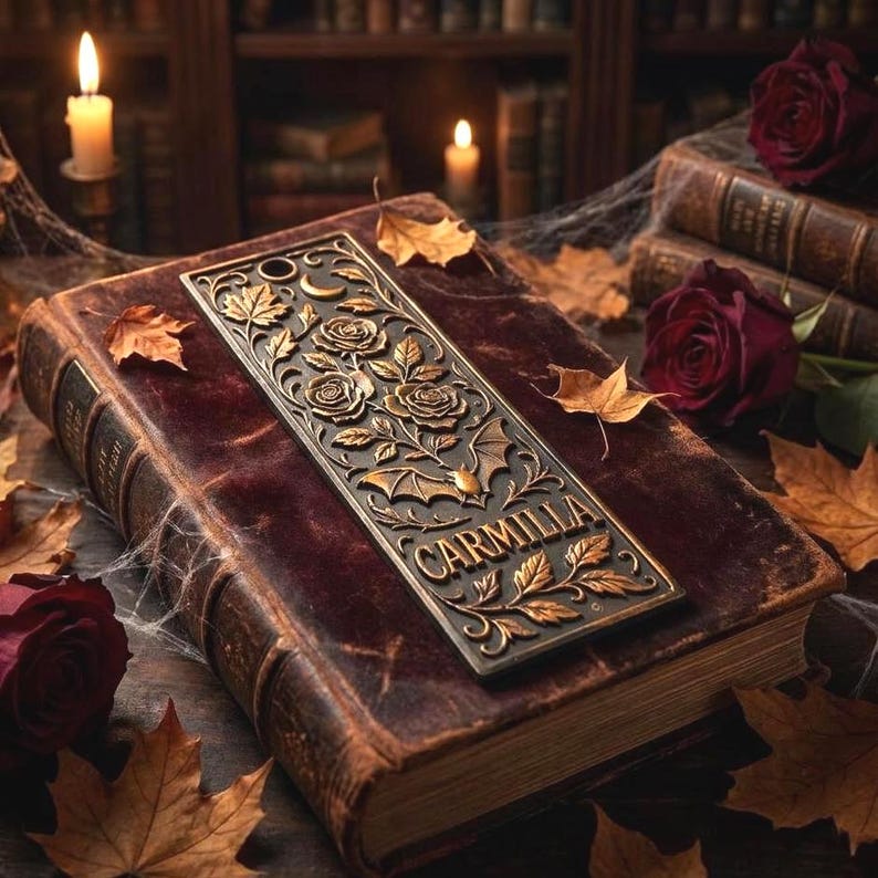 May include: A bronze bookmark with floral and bat designs, and the word "CARMILLA" is placed on a vintage book with a burgundy cover. The scene includes autumn leaves, roses, and lit candles, creating a gothic aesthetic.