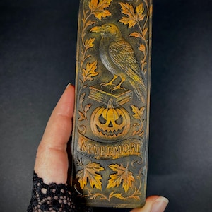 May include: A rectangular, dark brown bookmark with a carved raven perched atop a book and a jack-o'-lantern. The bookmark features the word "NEVERMORE" on a banner, surrounded by autumn leaves. The raven is detailed with yellow and brown accents.