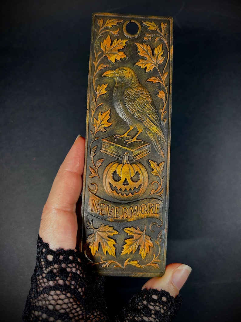 May include: A rectangular, dark brown bookmark with a carved raven perched atop a book and a jack-o'-lantern. The bookmark features the word "NEVERMORE" on a banner, surrounded by autumn leaves. The raven is detailed with yellow and brown accents.