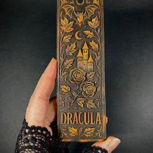 May include: A rectangular, dark brown bookmark with the word "DRACULA" in gold lettering. The bookmark features a carved design of a castle, roses, leaves, a bat, and a crescent moon. The overall theme is gothic and literary.