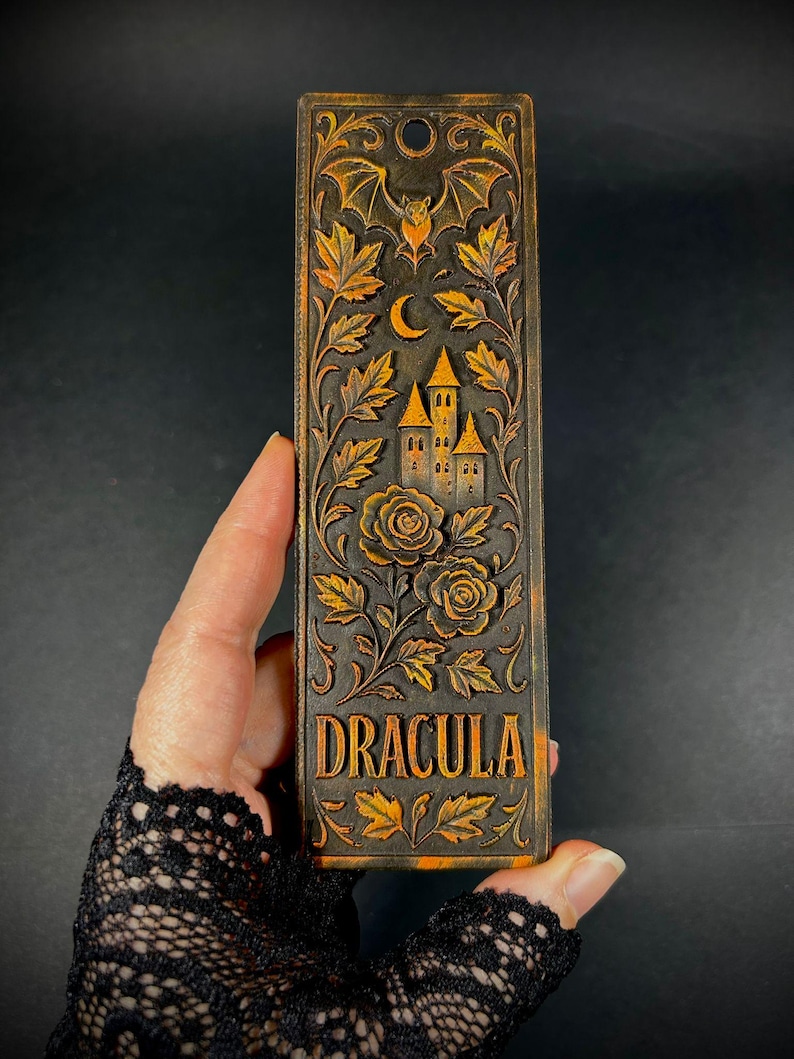 May include: A rectangular, dark brown bookmark with the word "DRACULA" in gold lettering. The bookmark features a carved design of a castle, roses, leaves, a bat, and a crescent moon. The overall theme is gothic and literary.