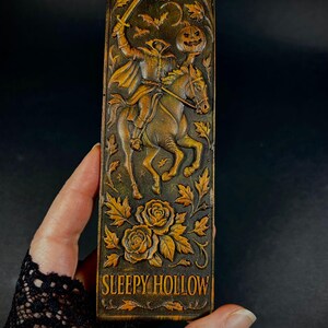 May include: A rectangular, brown bookmark with a carved design featuring the Headless Horseman riding a horse, holding a sword and a jack-o'-lantern. The text "SLEEPY HOLLOW" is at the bottom, with floral and bat accents.