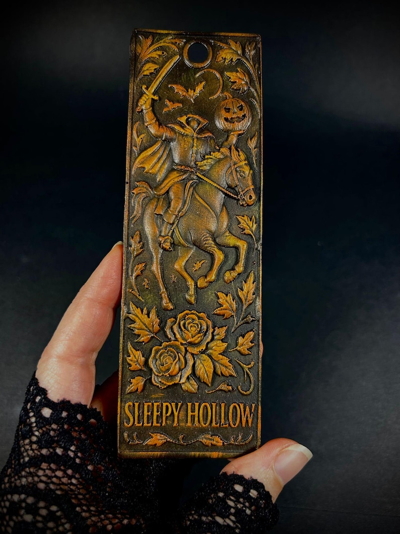 May include: A rectangular, brown bookmark with a carved design featuring the Headless Horseman riding a horse, holding a sword and a jack-o'-lantern. The text "SLEEPY HOLLOW" is at the bottom, with floral and bat accents.