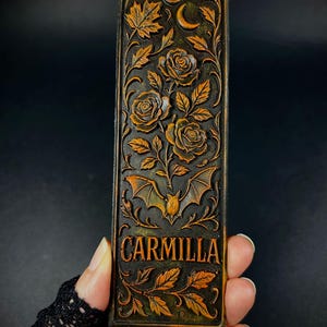May include: A rectangular, dark brown bookmark with an embossed design featuring roses, leaves, a crescent moon, and a bat. The word "CARMILLA" is displayed in gold lettering. The bookmark has a hole at the top for a tassel.