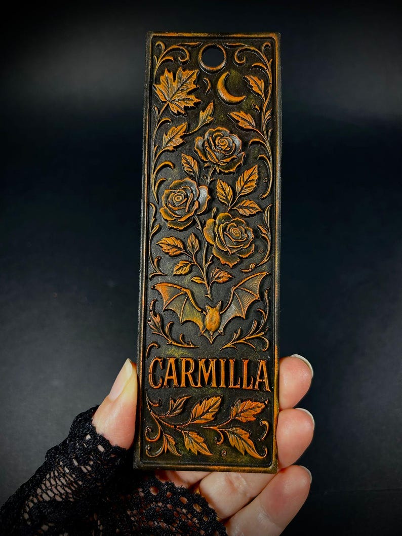 May include: A rectangular, dark brown bookmark with an embossed design featuring roses, leaves, a crescent moon, and a bat. The word "CARMILLA" is displayed in gold lettering. The bookmark has a hole at the top for a tassel.