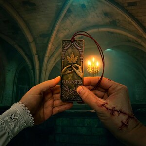 May include: A bronze-colored bookmark with a maple leaf design and the word "Frankenstein". The bookmark is held by two hands, one with stitches. A red cord is attached to the top. The background features a dimly lit room with arched ceilings.
