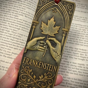 May include: Bronze-colored metal bookmark featuring the word "FRANKENSTEIN" and an illustration of a leaf held by two hands. The bookmark has an arched top and a red cord for easy placement in a book.